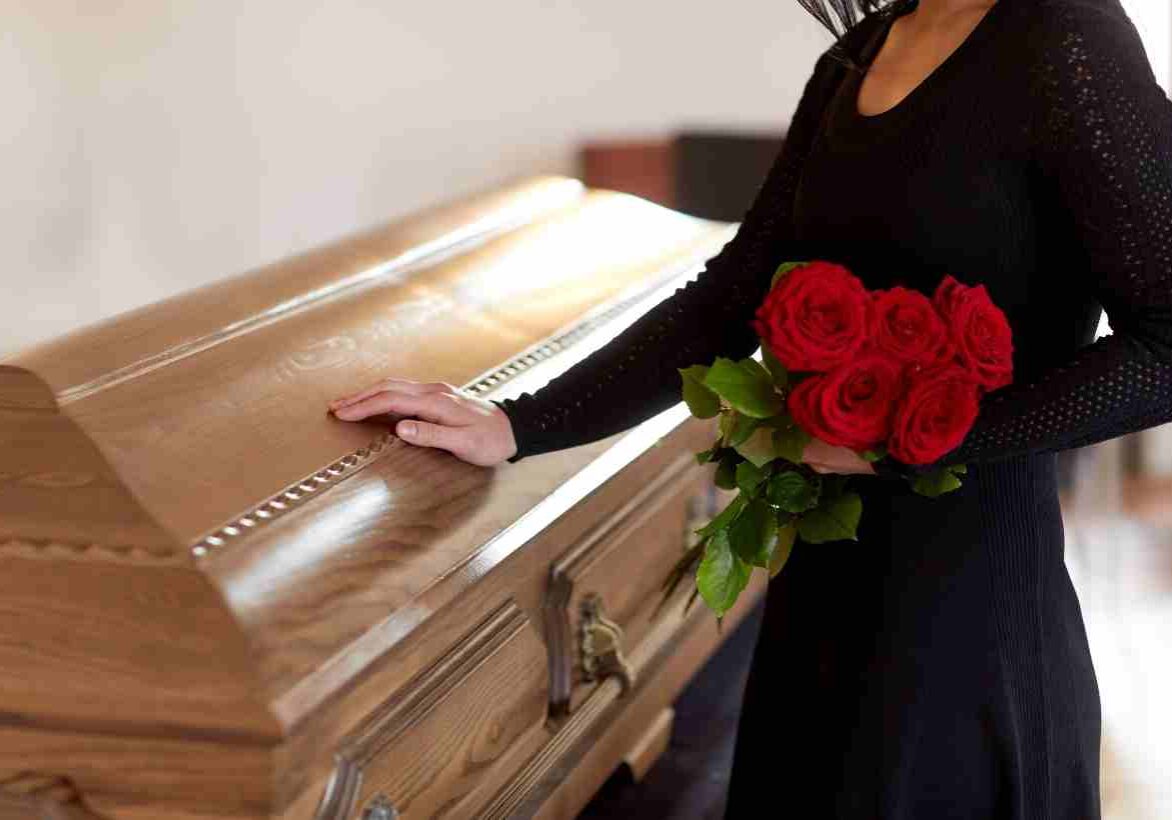 woman in black dress holding flowers with hand on coffin